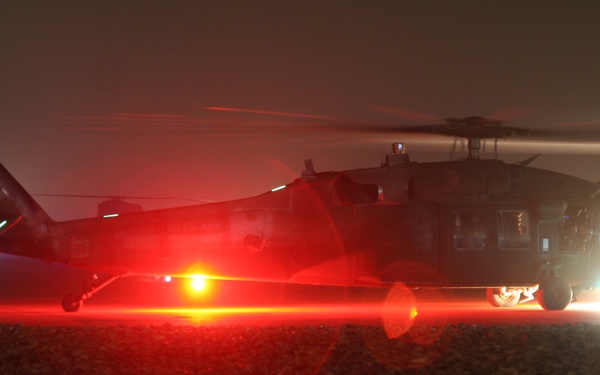 A military helicopter, identifiable as a UH-60 Black Hawk (tail number 060), is photographed at night on a gravel landing zone with rotors spinning. The scene is illuminated by the aircraft's navigation and landing lights, including a prominent red belly light and a bright white searchlight, creating dramatic light flares. The low-visibility, foggy nighttime conditions suggest an active operational or training environment.