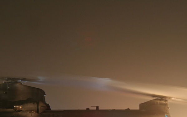 A large tandem-rotor helicopter, consistent with a CH-47 Chinook, is photographed at night on what appears to be a forward operating base or military airfield. The rotors are spinning, indicated by motion blur, and the aircraft appears to be preparing for departure or has just landed. The scene is illuminated by colored navigation lights and ground lighting in hazy/dusty atmospheric conditions.