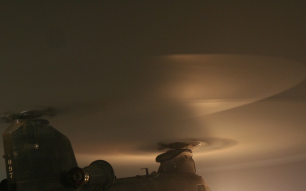 A large tandem-rotor military helicopter, identifiable as a CH-47 Chinook, is photographed at night on what appears to be a military airfield or flight line. The rotors are spinning, visible as motion-blurred arcs above the aircraft, suggesting the helicopter is either preparing for takeoff or has just landed. Ambient lighting from ground sources and navigation lights illuminate the scene through fog or haze.