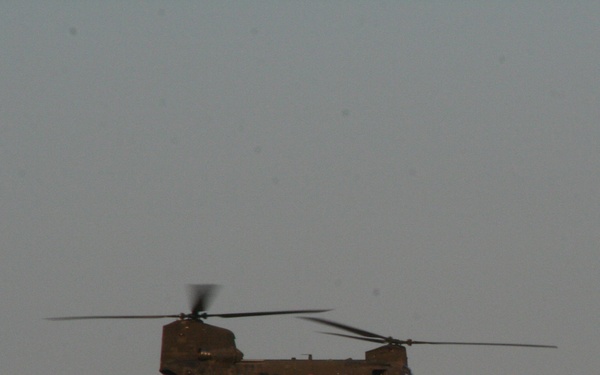 A tandem-rotor military helicopter, consistent with a CH-47 Chinook configuration, is photographed in flight against an overcast gray sky. The helicopter is carrying a sling load — a small cargo item suspended beneath the fuselage on a cable. The image appears to have been taken at dusk or dawn, resulting in low ambient light and muted tones.