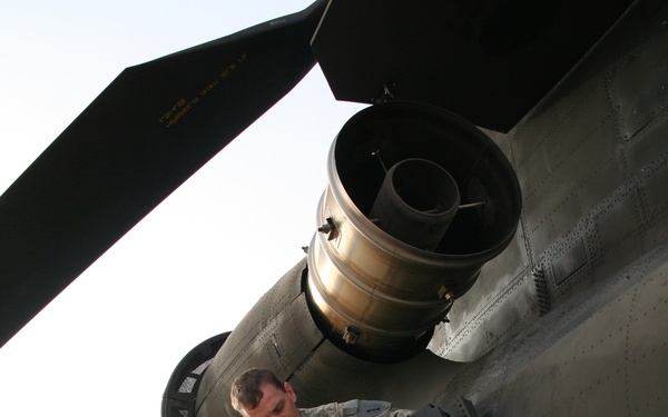 A U.S. Army soldier in ACU (Army Combat Uniform) camouflage is performing maintenance or inspection work on the exterior of a large military helicopter, identified by its twin-rotor configuration as a CH-47 Chinook. The soldier is standing on a step or foothold on the fuselage while handling a black equipment box or avionics component mounted to the aircraft's side. The setting appears to be a forward operating base or airfield in an arid environment.