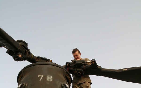 A military technician or mechanic is standing atop a large tandem-rotor helicopter, appearing to inspect or perform maintenance on the rotor head assembly. The aircraft is olive drab/dark green in color and displays the number '78' on the rotor pylon and a red '1' on the nose section. A caution label reading 'FRAGILE AREA DO NOT PUSH' is visible on the fuselage.