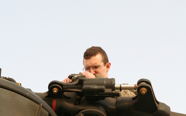 A military personnel wearing US Army ACU (Army Combat Uniform) is standing on top of a military helicopter, inspecting or working on the rotor hub assembly. The photo is taken from a low angle looking upward, showing the rotor head mechanism in the foreground. The aircraft appears to be an olive drab military helicopter, likely a CH-47 Chinook or similar large rotary-wing aircraft based on the fuselage construction.
