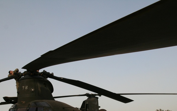 A large military tandem-rotor helicopter, identified as a CH-47 Chinook marked with the number '78', is parked on a concrete flight line at what appears to be a forward operating base or military airfield. At least one person is visible on top of the fuselage, likely performing maintenance or inspection. A second Chinook helicopter is visible in the background, along with base infrastructure including hangars and concrete barriers.
