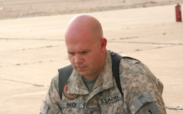 A U.S. Army soldier identified by name tape as 'BECK' is seated on a concrete surface operating a Dell laptop computer placed on top of a hard-shell equipment case. He is wearing Army Combat Uniform (ACU) in the Universal Camouflage Pattern with a '1' patch visible on his right sleeve and a unit patch on his left shoulder. A fire extinguisher is visible in the background, suggesting a military installation or airfield environment.