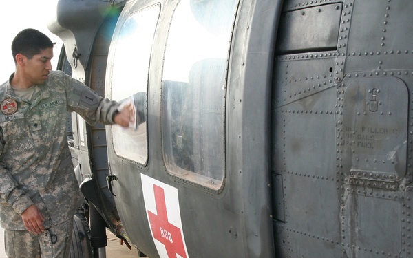 A U.S. Army soldier in ACU (Army Combat Uniform) is inspecting or performing a pre-flight check on a military medical evacuation (MEDEVAC) helicopter, identifiable by the prominent Red Cross symbol on its fuselage. The aircraft appears to be a UH-60 Black Hawk configured for medical evacuation duties, parked on a tarmac in what appears to be an arid environment. The soldier's name tape reads 'MOYA' and he is reaching toward the helicopter's window area.