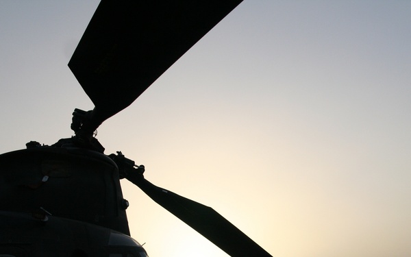 A large tandem-rotor helicopter, consistent with a Boeing CH-47 Chinook, is photographed in silhouette against a hazy sunset sky. The aircraft is parked on a flat, arid desert terrain, suggesting a forward operating base or austere landing zone. The backlit composition renders nearly all detail in deep shadow, emphasizing the aircraft's distinctive rotor and fuselage profile.