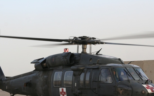 A dark olive/black UH-60 Black Hawk helicopter configured as a MEDEVAC (medical evacuation) aircraft is on a tarmac, with its rotors spinning. The helicopter displays prominent red cross markings on its fuselage, indicating its medical role under the Geneva Convention. A soldier in combat uniform and flight helmet is standing near the aircraft, connected by a ground communication cord.