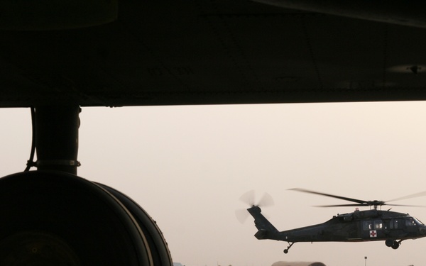A military medical evacuation (MEDEVAC) helicopter, identifiable by the red cross emblem on its fuselage, is captured hovering or landing on a military airfield. The photo is taken from beneath a large aircraft, with its landing gear wheel prominently silhouetted in the foreground. The background reveals hardened aircraft shelters and base infrastructure consistent with a forward operating base in an arid environment.