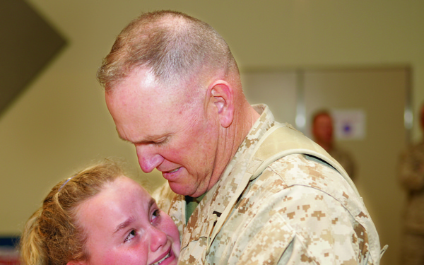 A U.S. Marines service member in desert MARPAT camouflage uniform embraces a young girl, approximately 8-12 years old, who is wearing a bright pink shirt. The girl appears to be crying tears of joy or emotion while smiling, suggesting a reunion scenario. The setting appears to be an indoor facility, likely a military base or gymnasium.