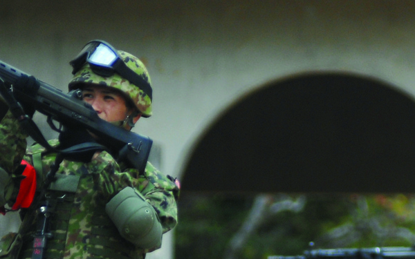 Two soldiers in camouflage uniforms are conducting what appears to be a military training exercise near a building structure. The foreground soldier is holding a rifle raised upward while wearing full tactical gear including helmet with goggles and knee pads. A second soldier is visible in the background aiming a rifle in a combat-ready stance.