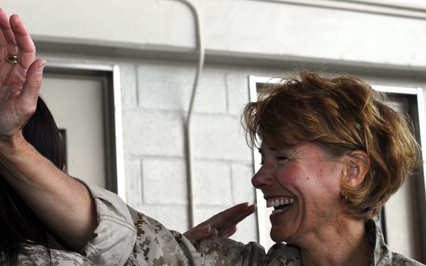 Two individuals wearing USMC MARPAT desert camouflage uniforms are engaged in a warm, joyful interaction. The woman facing the camera is laughing broadly while embracing the other person, who has one hand raised. The setting appears to be an indoor military facility with concrete block walls.