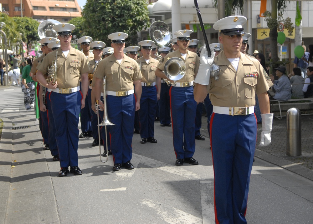 A United States Marine Corps marching band is performing in a parade on a city street. The Marines are dressed in their distinctive Service Alpha uniforms with khaki tops, blue trousers with red stripes, and white covers. The setting appears to be in Japan, as indicated by visible Japanese kanji signage in the background.