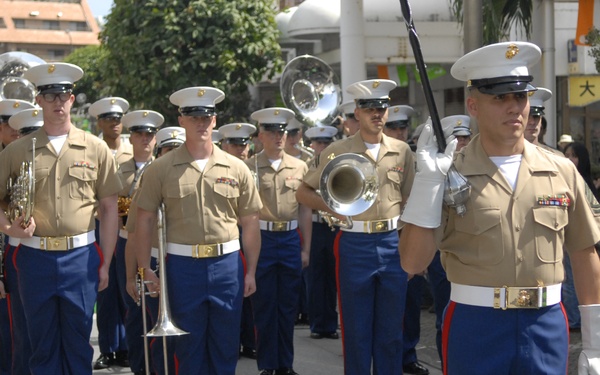 A United States Marine Corps marching band is performing in a parade on a city street. The Marines are dressed in their distinctive Service Alpha uniforms with khaki tops, blue trousers with red stripes, and white covers. The setting appears to be in Japan, as indicated by visible Japanese kanji signage in the background.