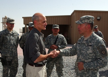 A civilian in a dark shirt and khaki cargo pants is shaking hands with a U.S. Army officer in ACU (Army Combat Uniform) at what appears to be a forward operating base or military installation. Several uniformed soldiers stand in the background observing the greeting. The setting appears to be an arid, desert environment based on the terrain and building construction.