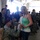 A U.S. Army soldier wearing ACU (Army Combat Uniform) and a maroon beret is kneeling on one knee, appearing to propose to a young blonde woman in a green strapless top and jeans. The scene takes place inside what appears to be a military hangar or assembly area, surrounded by other soldiers and civilians. The crowd of onlookers in the background suggests this is a military homecoming or deployment event.