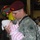 A U.S. Army soldier wearing an 82nd Airborne Division patch and maroon beret tenderly holds a newborn baby dressed in pink and white striped clothing. The setting appears to be a military homecoming event, evidenced by 'Welcome Back' balloons visible in the background. The soldier is in full ACU (Army Combat Uniform) with a rucksack, suggesting he has just returned from deployment.