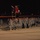 A large formation of U.S. Army soldiers in ACU (Army Combat Uniform) camouflage is marching across an airfield tarmac at night. Two guidon/color bearers are visible near the front carrying unit flags, one of which appears to display the number '2'. The scene is consistent with a unit deployment ceremony, homecoming, or formal military movement at a military airfield.
