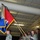 A U.S. Army military ceremony taking place inside a hangar or large indoor facility. Soldiers in ACU (Army Combat Uniform) are participating in a color guard or unit ceremony, with personnel holding guidons/flags and rifles. The prominent flag displayed is red and blue with the number '2' visible, consistent with a brigade or battalion colors presentation.