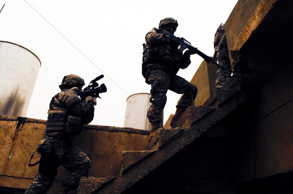 Three soldiers in military combat uniforms and helmets are conducting a tactical operation on what appears to be a rooftop or elevated concrete structure. One soldier on the left is in a crouched position aiming a weapon with an optical attachment, while two others are ascending concrete stairs. The image is taken from a low angle in overcast lighting conditions, creating a dramatic silhouette effect.