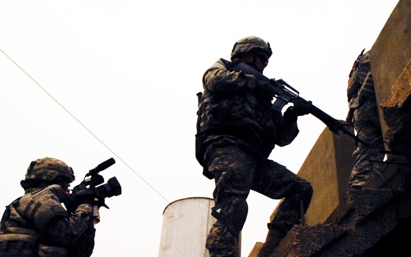 Three soldiers in military combat uniforms and helmets are conducting a tactical operation on what appears to be a rooftop or elevated concrete structure. One soldier on the left is in a crouched position aiming a weapon with an optical attachment, while two others are ascending concrete stairs. The image is taken from a low angle in overcast lighting conditions, creating a dramatic silhouette effect.