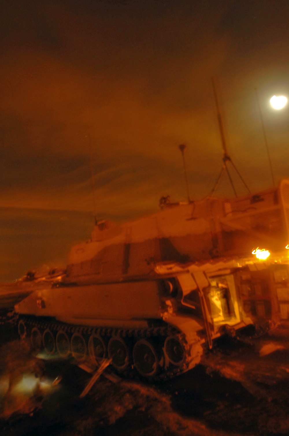 A military armored vehicle, likely a tank or self-propelled artillery piece, photographed at night under orange-tinted artificial lighting. The vehicle appears stationary and is viewed from a low angle, showing its tracked undercarriage and turret. Multiple antennas are visible extending from the vehicle, and background lights suggest a populated or base area nearby.