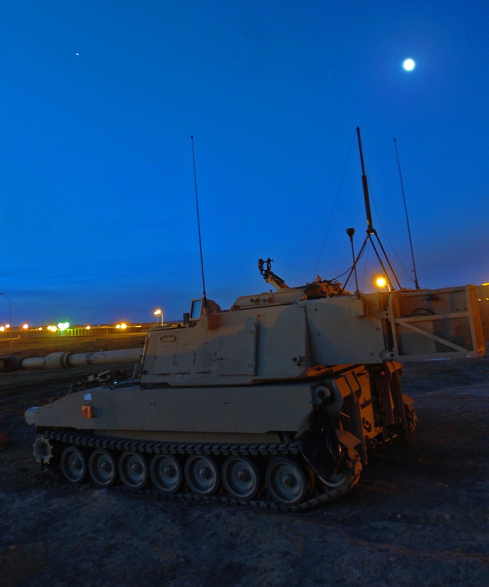 A self-propelled howitzer, consistent with the M109 Paladin series, is photographed at dusk or dawn in a desert/arid military installation. The vehicle is painted in a tan/desert camouflage scheme and is stationary on open ground. The moon is visible in the deep blue sky, and base lighting illuminates the background.