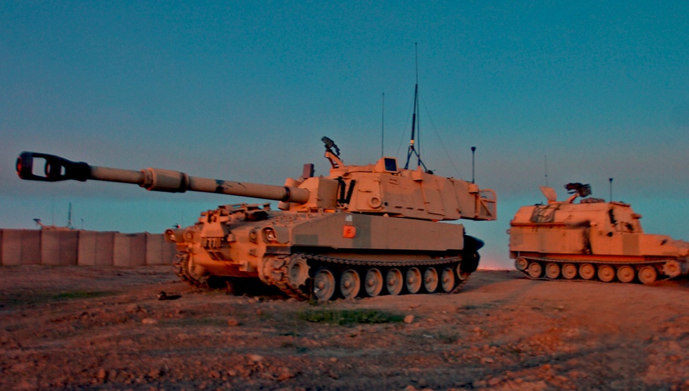 The image shows two self-propelled artillery vehicles parked on arid, flat terrain during twilight or dusk. The foreground vehicle appears to be an M109 Paladin 155mm self-propelled howitzer in desert tan camouflage, with a second similar tracked vehicle visible in the background. The scene is illuminated by warm orange ambient light from the setting sun contrasting with a blue sky.