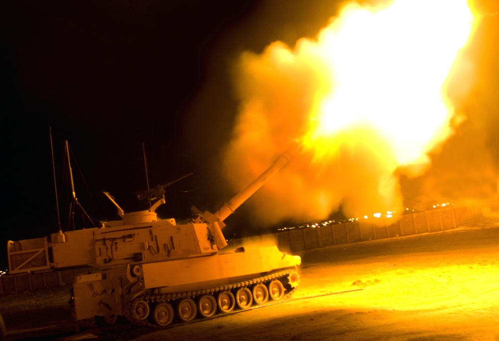 A self-propelled artillery piece is captured mid-fire during nighttime operations, producing a massive muzzle blast with intense white and orange flames. The vehicle appears to be an M109 Paladin or similar tracked self-propelled howitzer, illuminated dramatically by its own discharge. The surrounding terrain appears to be arid/desert ground, with what appears to be a barrier wall or HESCO barriers visible in the background.