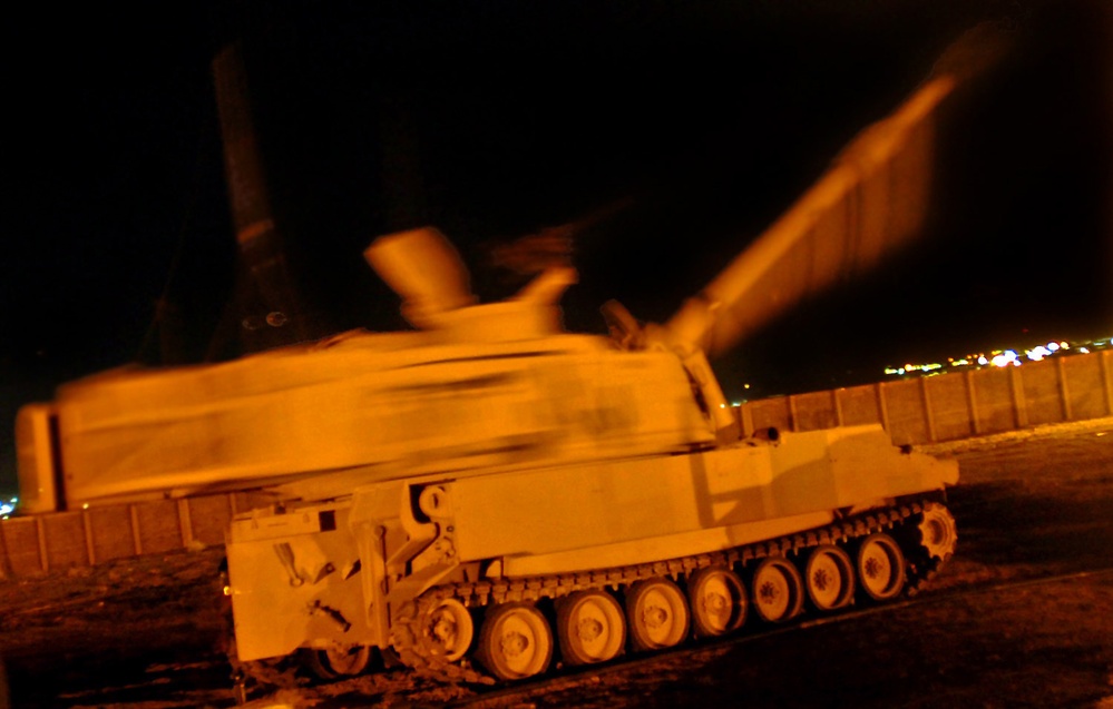 A tracked military self-propelled artillery vehicle is photographed at night with significant motion blur, suggesting the turret or gun barrel is rotating or the vehicle is in motion. The scene is illuminated by an orange artificial light source, casting a warm glow over the vehicle. Concrete barriers and distant city lights are visible in the background.