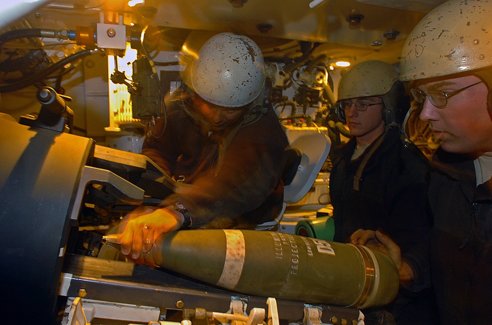 Three military personnel wearing protective helmets are working inside what appears to be a naval gun turret or artillery compartment. They are handling a large artillery round or projectile, with text visible on the shell reading 'ILLUMINATING PROJECTILE'. The interior space is cramped and filled with mechanical equipment, illuminated by warm artificial lighting.