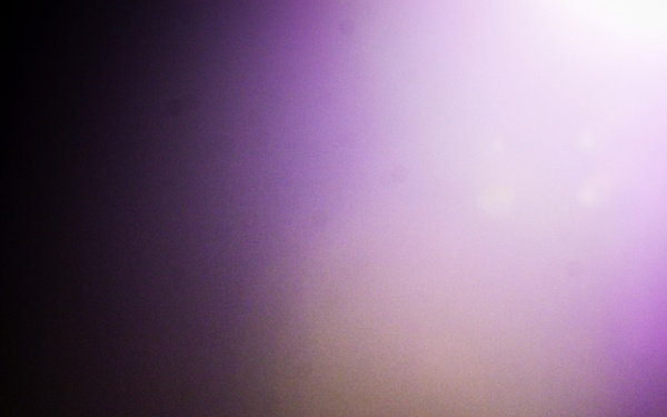 A low-light photograph showing silhouetted figures in the foreground against a dramatic purple and white stage light beam cutting through haze or smoke. The scene appears to be a live music concert or performance event, captured from within or near the audience. The strong backlighting creates deep shadows, rendering most subjects as dark silhouettes.