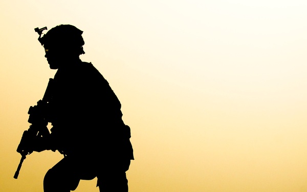 A silhouetted soldier in full combat gear is kneeling on an elevated ridge against a warm golden-yellow sky, likely at sunrise or sunset. The figure is rendered entirely in dark silhouette, emphasizing the dramatic contrast between the subject and the glowing background. The composition conveys a sense of vigilance and solitude in a desert or arid environment.