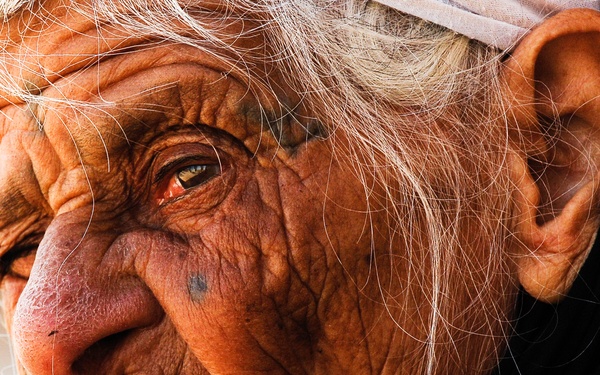 A close-up portrait of an extremely elderly person with deeply weathered and heavily wrinkled skin, white hair, and a contemplative expression. The subject is wearing dark clothing and appears to have a small mark or mole on their cheek. The photograph is taken with dramatic side lighting that emphasizes the texture and depth of the facial features.