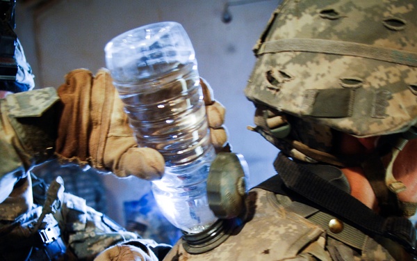 Two soldiers in US Army digital camouflage (ACU pattern) uniforms and helmets are visible in a low-light environment. One soldier is holding up a clear plastic water bottle, which is illuminated and appears to be the primary light source in the scene. The image appears to capture a field or operational setting where soldiers are managing water supplies.
