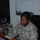 A U.S. Army soldier named Morris, wearing Army Combat Uniform (ACU) with a Staff Sergeant rank insignia, is seated at a desk working on a laptop computer. The setting appears to be a military office or operations center, with a whiteboard containing notes visible in the background. Two laptops are present on the desk along with other office equipment.