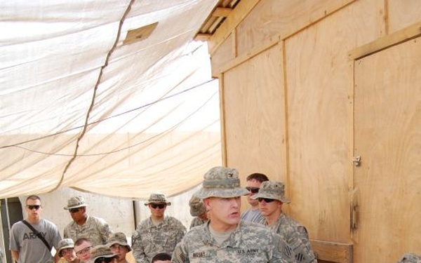A U.S. Army officer or senior NCO is addressing a group of soldiers gathered at what appears to be a forward operating base or military camp. The central figure is standing on a wooden pallet platform, speaking to the assembled troops who are seated or standing around him. The setting features improvised structures including plywood buildings and shade canopies typical of deployed military environments.