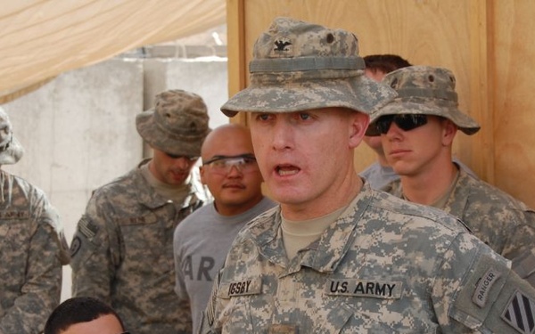 A group of U.S. Army soldiers in ACU (Army Combat Uniform) camouflage are gathered in what appears to be a forward operating base or outdoor military compound. The central figure, whose name tape reads 'UCSBY' or similar, is speaking or briefing others while wearing a boonie hat with a colonel's eagle insignia. The setting appears to be a desert or arid environment based on the sandy tones and construction materials visible.