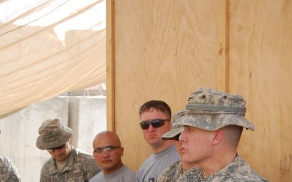 A group of U.S. Army soldiers gathered in what appears to be a forward operating base or construction area with plywood walls and a shade canopy overhead. One soldier in full ACU (Army Combat Uniform) with a patrol cap stands with arms crossed, appearing to address or brief the group. Other personnel are present in both ACUs and Army PT (physical training) uniforms.