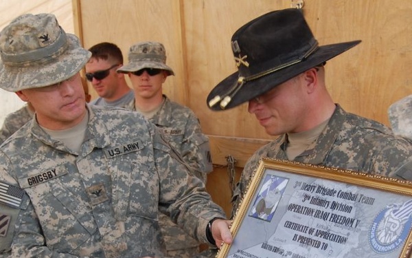 A U.S. Army ceremony in what appears to be a forward operating base, where a soldier named Grigsby is presenting or receiving a framed Certificate of Appreciation from the 3rd Heavy Brigade Combat Team, 3rd Infantry Division, Operation Iraqi Freedom V. Multiple soldiers in ACU (Army Combat Uniform) pattern camouflage are present, with one officer wearing a distinctive black cavalry Stetson hat. The setting appears to be an outdoor or semi-covered area with plywood walls.