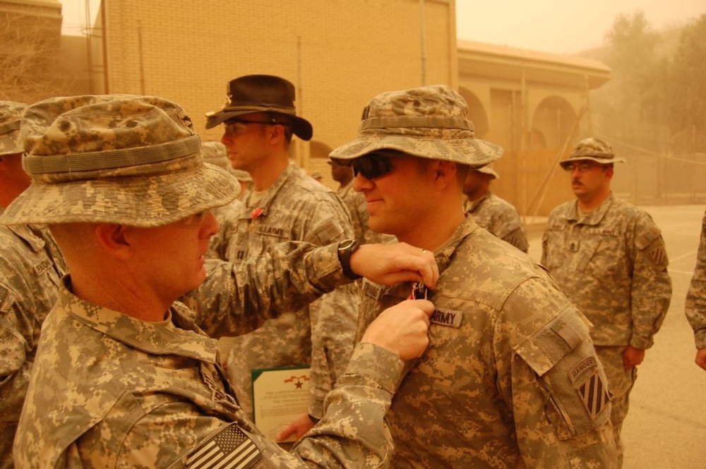A military award ceremony is taking place outdoors at what appears to be a forward operating base during dusty/sandy conditions. One soldier is pinning a medal or award onto another soldier's uniform chest. Multiple soldiers in US Army ACU (Army Combat Uniform) are present as witnesses.