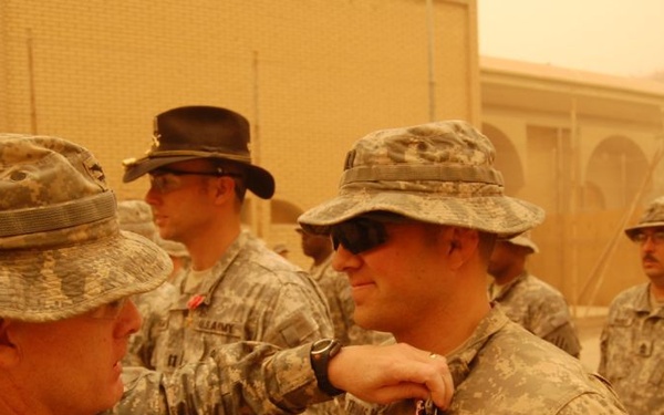 A military award ceremony is taking place outdoors at what appears to be a forward operating base during dusty/sandy conditions. One soldier is pinning a medal or award onto another soldier's uniform chest. Multiple soldiers in US Army ACU (Army Combat Uniform) are present as witnesses.