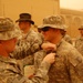 A military award ceremony is taking place outdoors at what appears to be a forward operating base during dusty/sandy conditions. One soldier is pinning a medal or award onto another soldier's uniform chest. Multiple soldiers in US Army ACU (Army Combat Uniform) are present as witnesses.