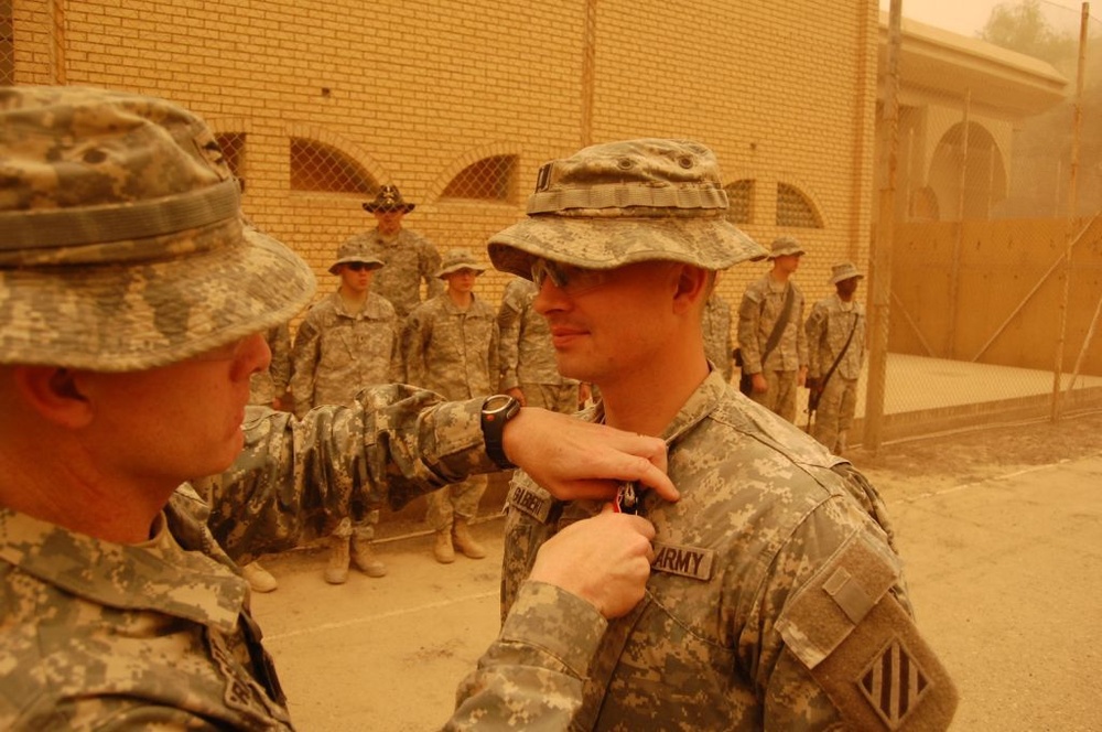 A military award or decoration ceremony is taking place outdoors in what appears to be a desert environment, likely Iraq or Afghanistan. A soldier is pinning a medal or badge onto the chest of another soldier wearing a boonie hat and Army combat uniform. Several other soldiers stand at attention in the background as witnesses to the ceremony.