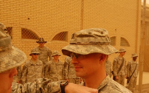 A military award or decoration ceremony is taking place outdoors in what appears to be a desert environment, likely Iraq or Afghanistan. A soldier is pinning a medal or badge onto the chest of another soldier wearing a boonie hat and Army combat uniform. Several other soldiers stand at attention in the background as witnesses to the ceremony.