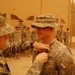 A military award or decoration ceremony is taking place outdoors in what appears to be a desert environment, likely Iraq or Afghanistan. A soldier is pinning a medal or badge onto the chest of another soldier wearing a boonie hat and Army combat uniform. Several other soldiers stand at attention in the background as witnesses to the ceremony.