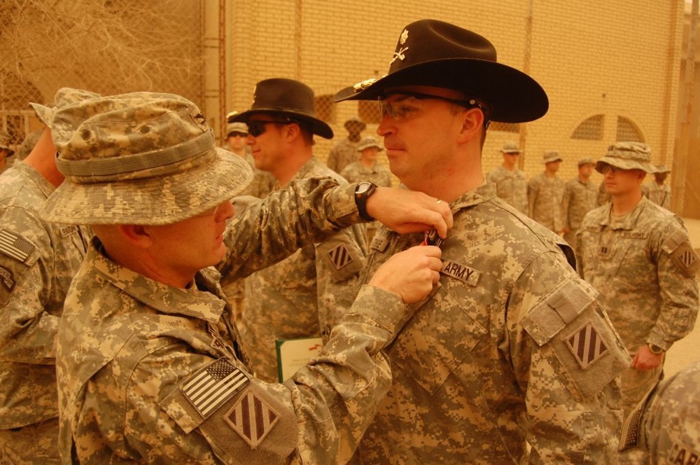 A military awards ceremony is taking place in what appears to be a forward operating base in a desert environment. A soldier wearing a distinctive black cavalry Stetson hat is receiving a medal or award being pinned to his uniform by another soldier. Multiple soldiers in ACU (Army Combat Uniform) are standing in formation in the background.