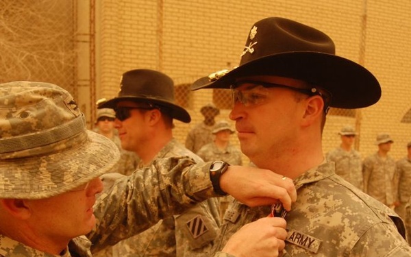 A military awards ceremony is taking place in what appears to be a forward operating base in a desert environment. A soldier wearing a distinctive black cavalry Stetson hat is receiving a medal or award being pinned to his uniform by another soldier. Multiple soldiers in ACU (Army Combat Uniform) are standing in formation in the background.