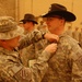 A military awards ceremony is taking place in what appears to be a forward operating base in a desert environment. A soldier wearing a distinctive black cavalry Stetson hat is receiving a medal or award being pinned to his uniform by another soldier. Multiple soldiers in ACU (Army Combat Uniform) are standing in formation in the background.