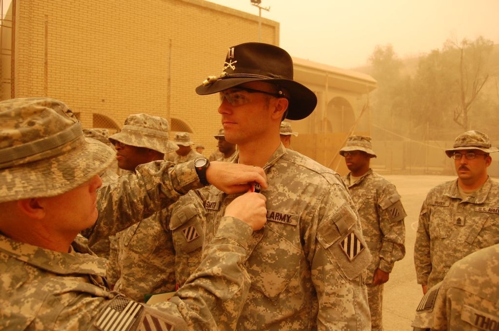 A U.S. Army soldier wearing a distinctive black cavalry Stetson hat is receiving a medal or award pinned to his uniform by a senior soldier during a military ceremony. The event appears to be taking place outdoors at a forward operating base in a desert environment, likely in Iraq or Afghanistan, under hazy, dust-filled conditions. Multiple soldiers in Army Combat Uniforms (ACU) are present as witnesses to the ceremony.